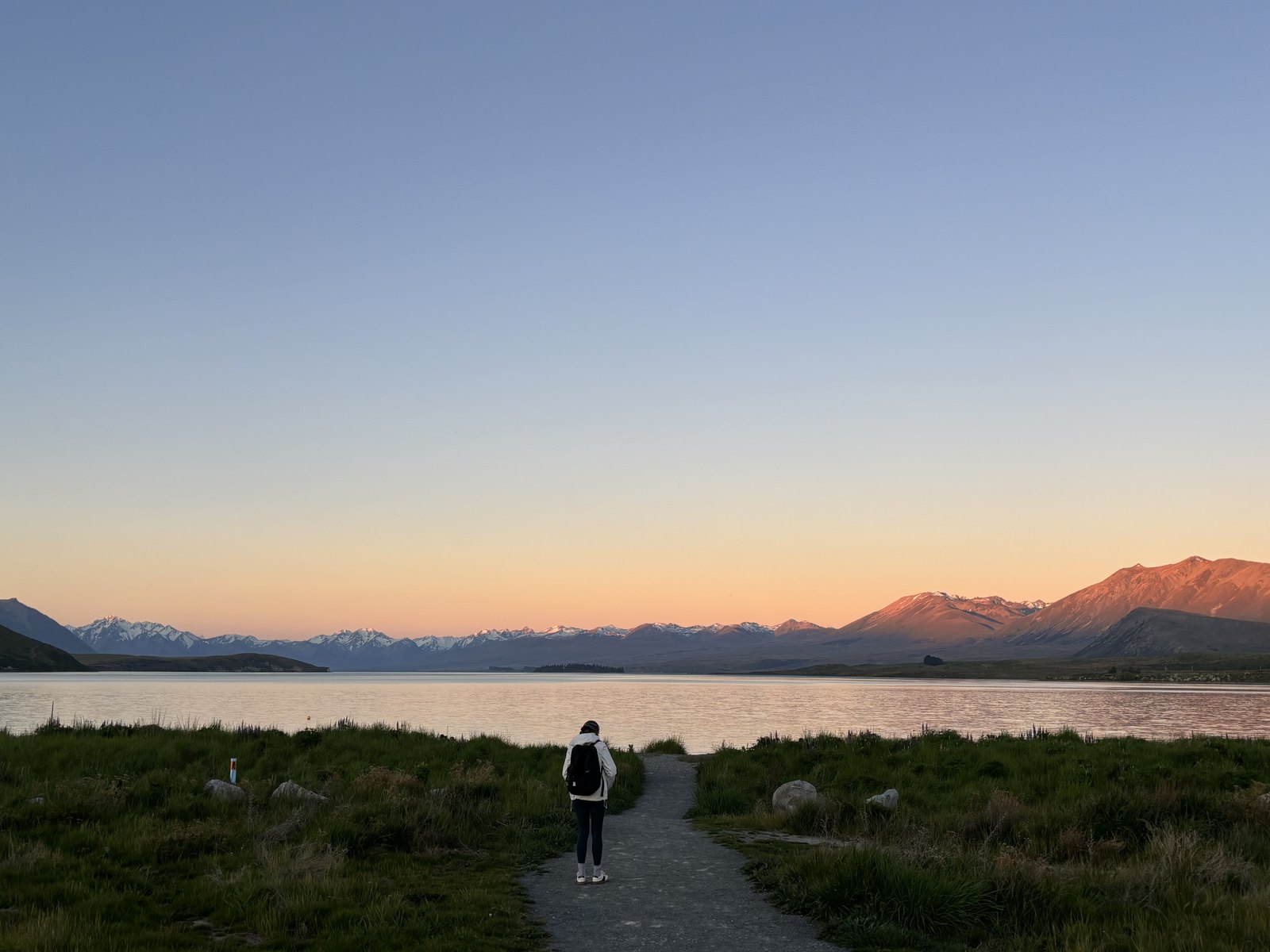 特卡波湖邊的小路上，一個人背着背囊，面向湖和晚霞中的遠山。