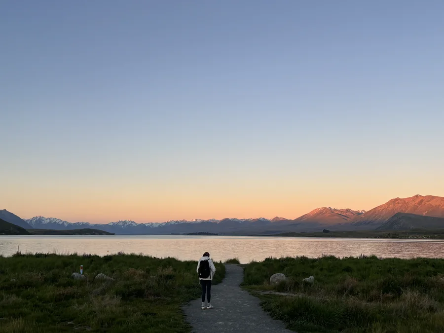 At dusk on Lake Tekapo, a traveller faces the lakeside mountains.